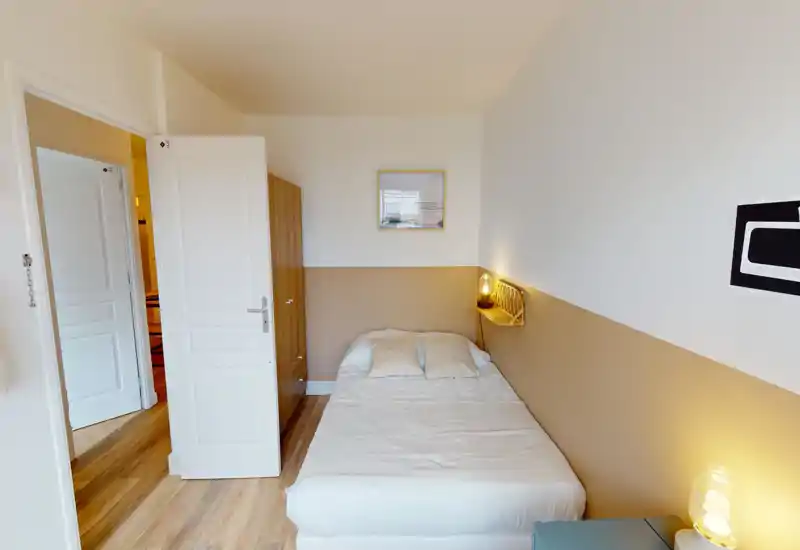 Sunny, compact bedroom with a neatly made single/double bed, warm bedside lighting and simple modern furnishings. Wooden floors and soft neutral colors create a cozy, inviting atmosphere ideal for short-term stays.