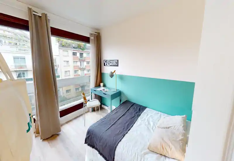 Bright, stylish single bedroom with a comfortable bed, a teal accent wall and a small desk by a large window letting in abundant natural light.