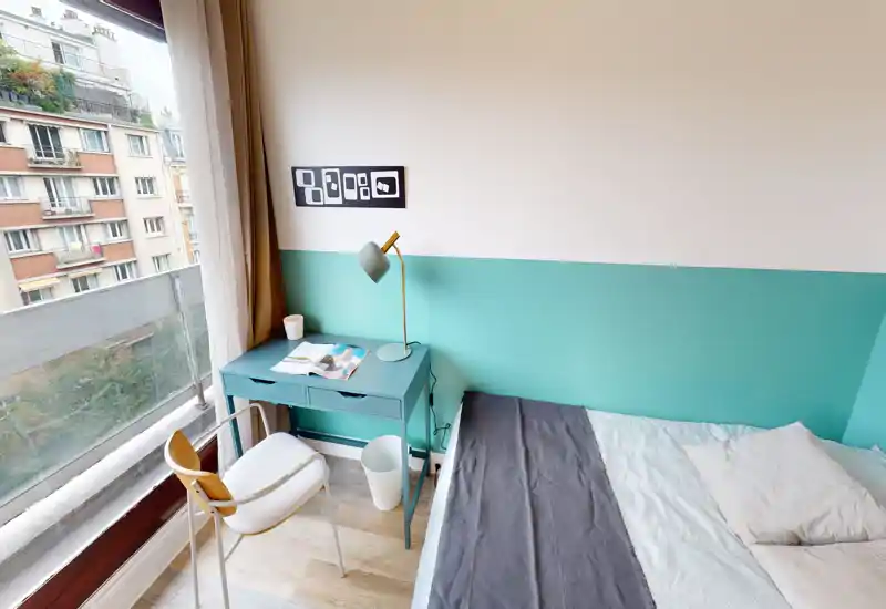 Bright, compact bedroom with a double bed, matching teal desk and lamp, and a large window opening onto city views — modern, tidy and well lit.