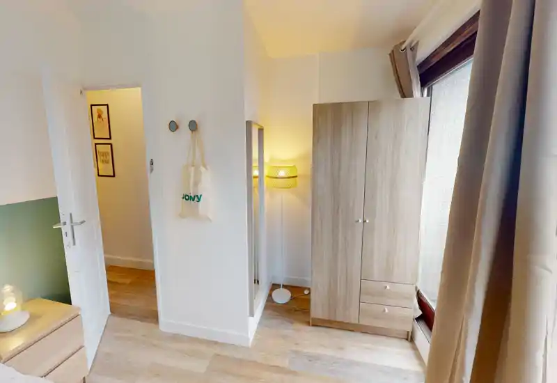 Small, well-lit bedroom corner with a wardrobe, bedside table and partial view of the bed; soft warm lamp light and neutral wood tones create a calm atmosphere.