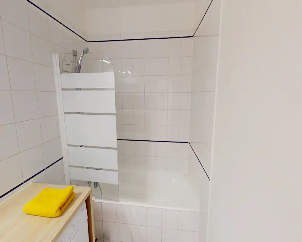 Salle de bains compacte et propre avec baignoire/douche carrelée, écran de douche en verre dépoli et petit plan de travail en bois avec serviettes pliées. Espace lumineux, net et fonctionnel, idéal pour de courts séjours.