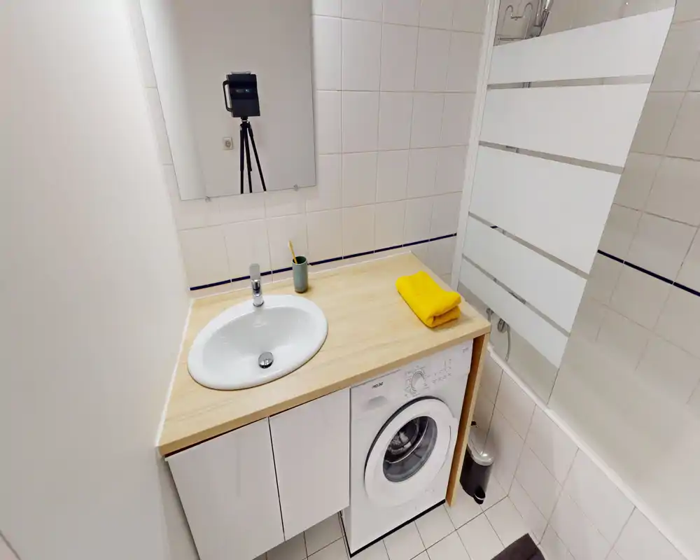 Salle de bain compacte avec lavabo, miroir et lave-linge intégré sous le plan de travail en bois ; carrelage blanc et pare-douche en verre pour un espace soigné.