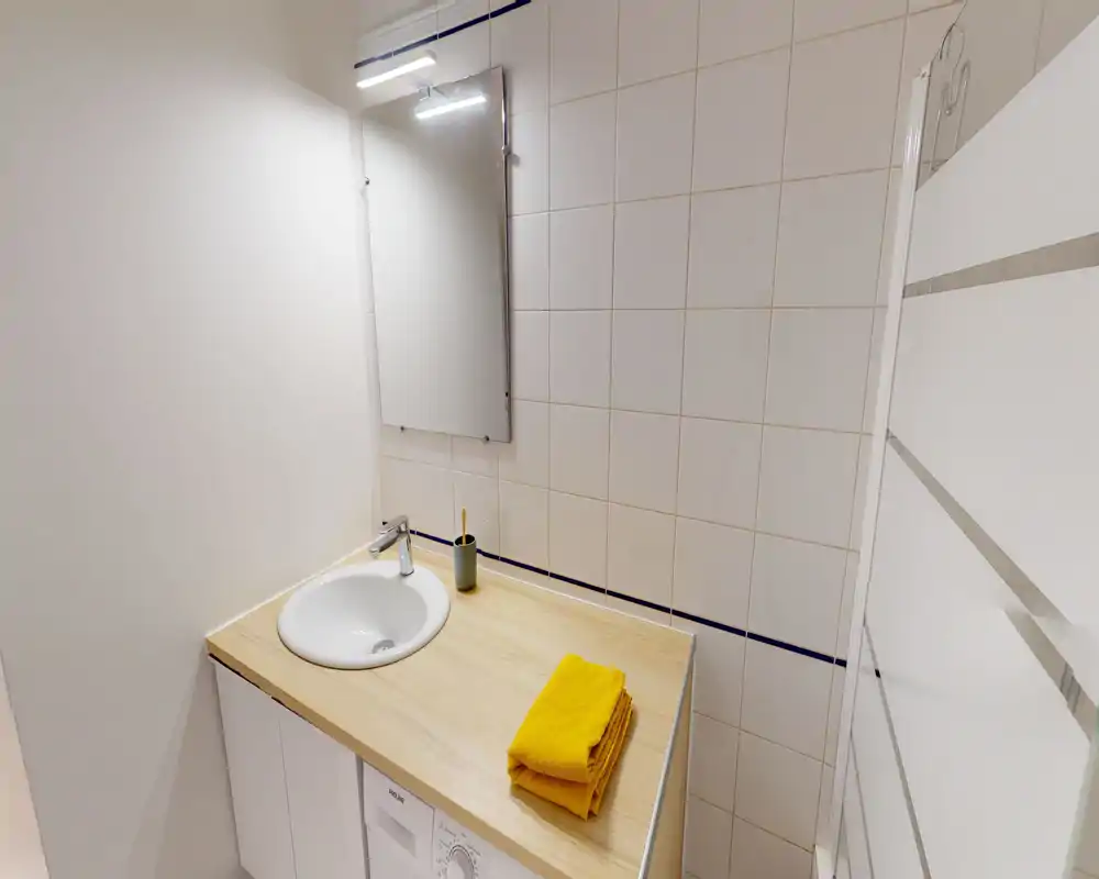 Espace lavabo de salle de bain moderne et compact avec plan de travail effet bois, vasque ronde, miroir et serviettes jaunes pliées — propre et lumineux.