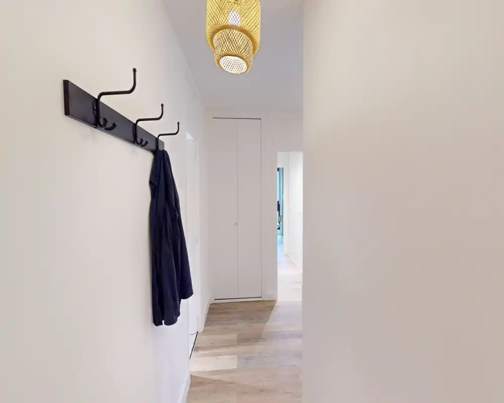 Entrée lumineuse et minimaliste avec patères murales et suspension, murs clairs et sol effet bois offrant un espace d’accueil net.