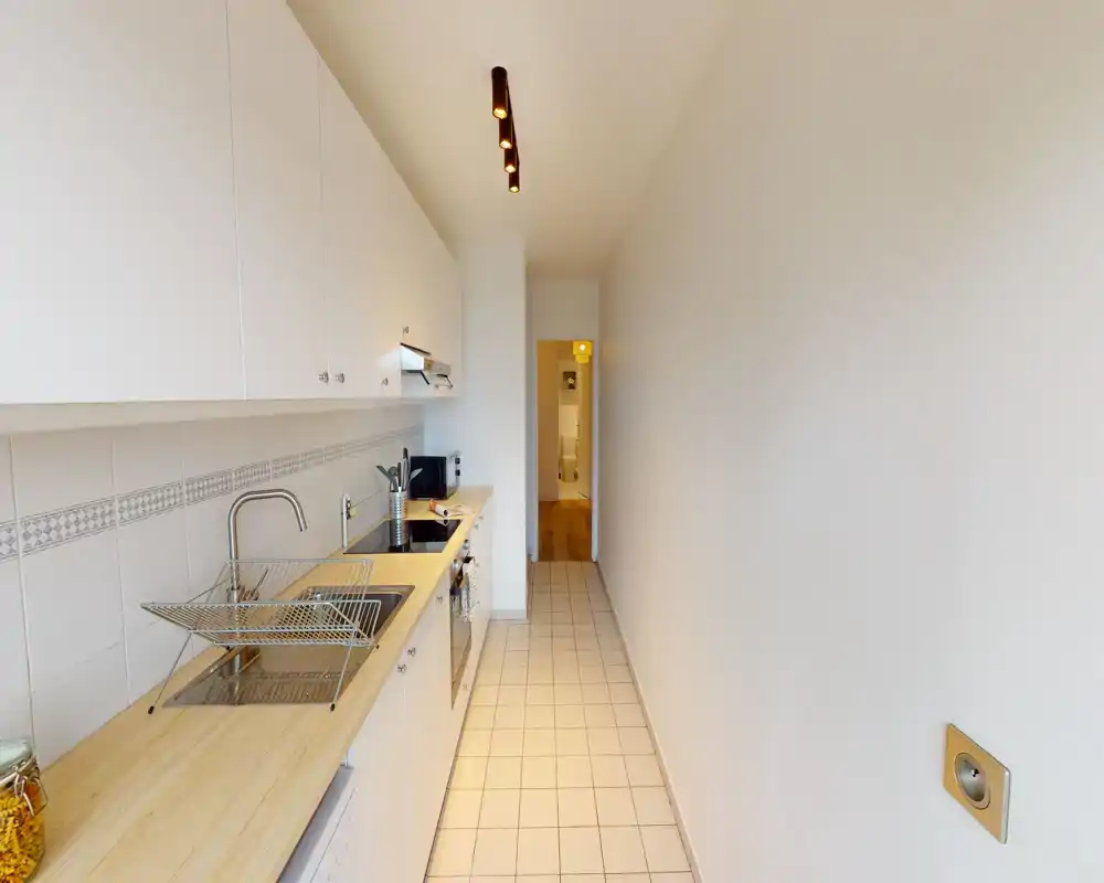 Cuisine en longueur lumineuse avec placards blancs, plan de travail en bois et sol carrelé — bien éclairée et rangée, parfaite pour un espace urbain compact.
