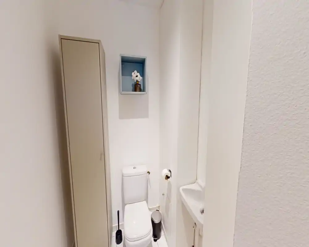 Petite salle de bains propre avec WC, lavabo compact et placard étroit ; murs blancs et accessoires soignés pour un espace fonctionnel.