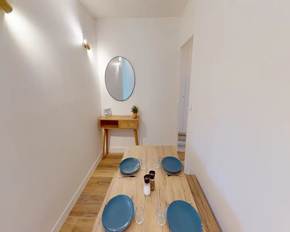 Coin repas lumineux et minimaliste avec une table en bois dressée pour quatre, assiettes bleues et une petite console avec miroir rond — style moderne et épuré, parfait pour les annonces.