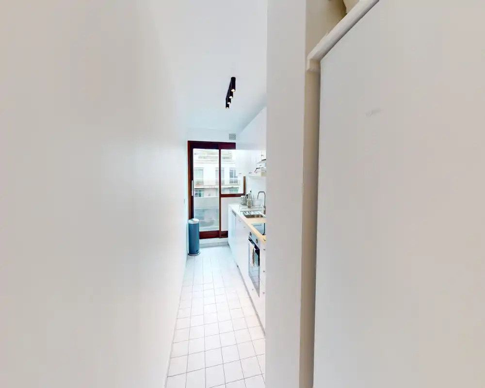 Cuisine en enfilade lumineuse avec meubles blancs, sol carrelé et une fenêtre/porte apportant la lumière naturelle ; agencement compact et fonctionnel.