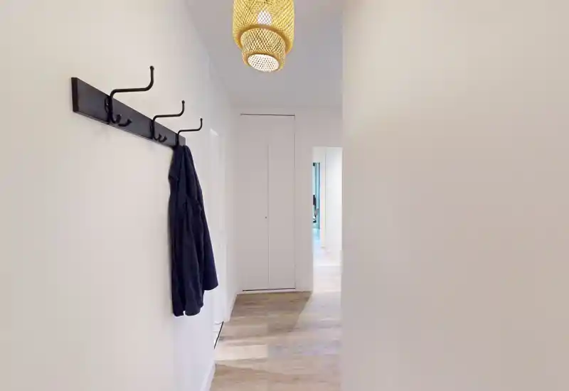 Bright, minimal entrance hallway with wall hooks and a pendant lamp, pale walls and wood-effect flooring creating a clean, welcoming arrival space.
