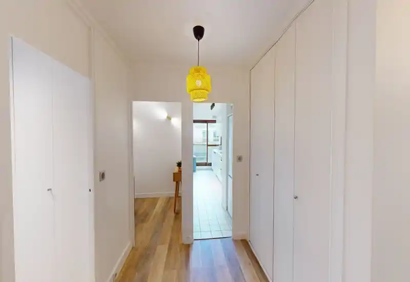 Bright, clean entrance hallway with built-in storage on both sides and a view into the kitchen. The wood-effect floor and central pendant lamp create a welcoming first impression.