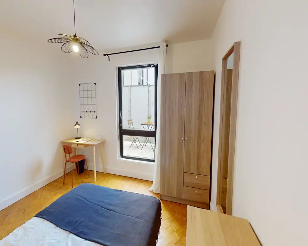 Chambre lumineuse et sobrement meublée avec un lit double, une armoire en bois, un bureau et une chaise près d'une fenêtre donnant sur une petite terrasse.