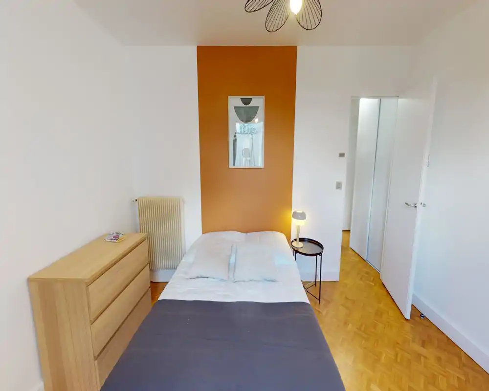 Chambre claire et soignée avec un lit double centré devant un panneau orange, lampe de chevet et commode en bois ; idéale pour une annonce valorisant un espace nuit chaleureux et lumineux.