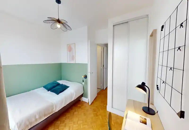 Bright single bedroom with a neatly made bed, green half-wall accent, wooden parquet floor and a small desk with lamp — ideal for a comfortable private stay.