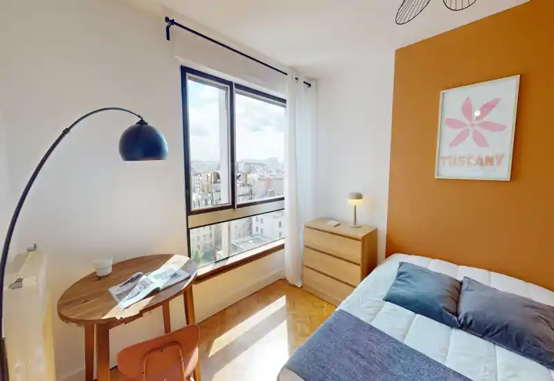 Camera da letto luminosa e ben arredata con letto confortevole, comò in legno e ampia finestra con vista sulla città. La parete colorata e i tessuti morbidi creano un'atmosfera accogliente, perfetta per annunci.