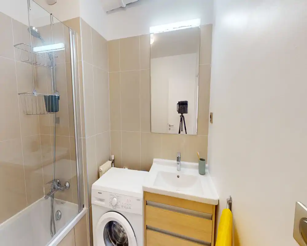 Modern compact bathroom with a bathtub-shower combination, sink with vanity and a front-loading washing machine tucked beside the basin. Neutral beige tiles and a wall mirror create a clean, functional feel.