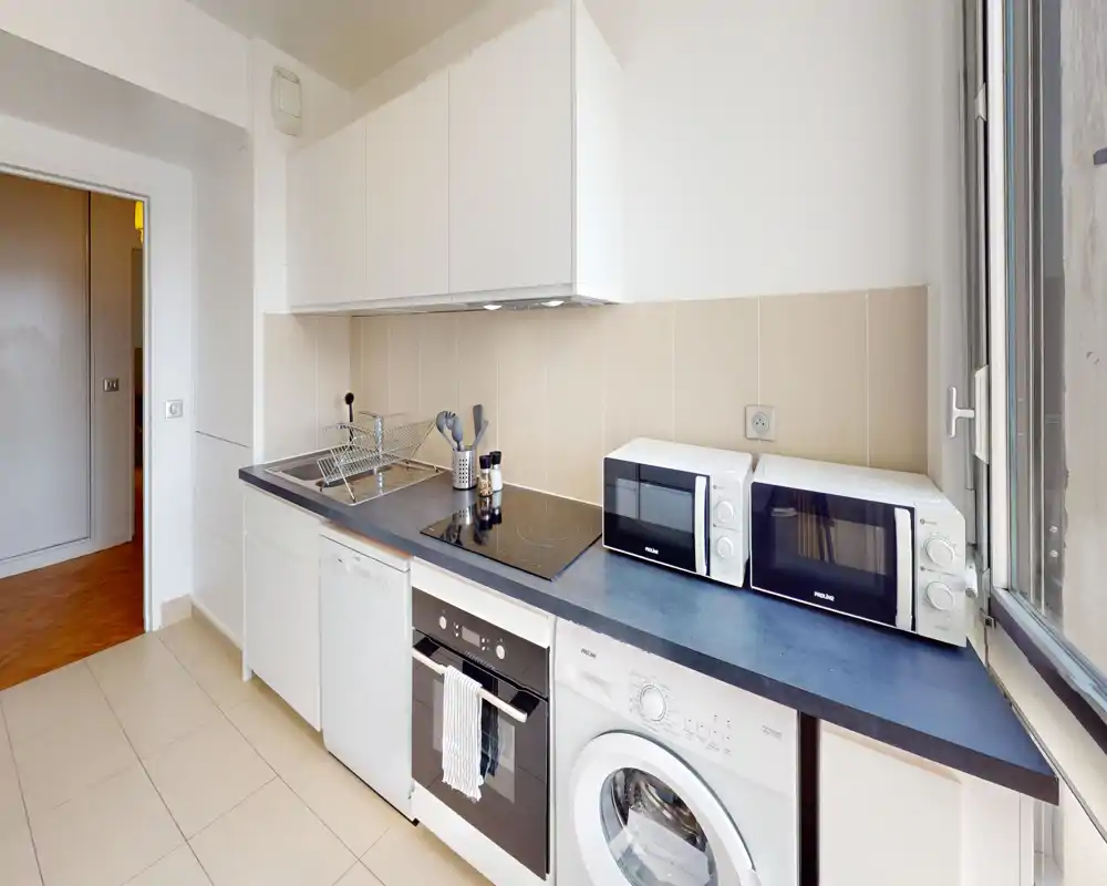 Bright, modern kitchen with integrated oven, induction hob, dishwasher and visible washing machine; two microwave ovens on the counter and a large window providing natural light.