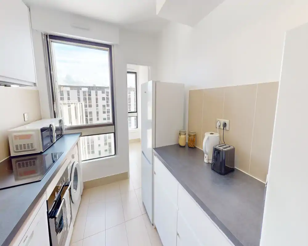Bright modern kitchen with large window, white cabinets, gray countertops and built-in appliances. The space is tidy and sunlit, featuring a fridge, microwave, toaster and kettle on the counter.