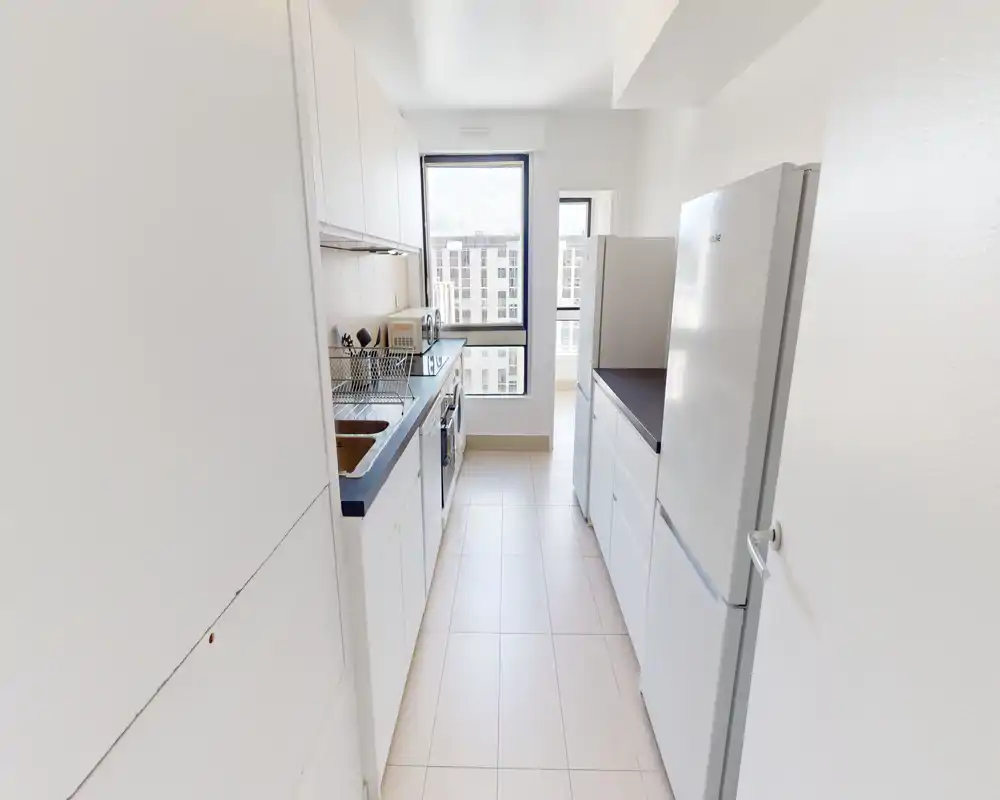 Bright, narrow galley kitchen with white cabinets, integrated sink, stove and a refrigerator, illuminated by a large window offering city views.