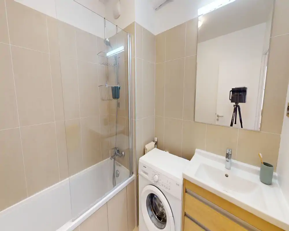 Bright, modern bathroom with a combined bathtub and shower, sink with vanity, mirror and a washing machine neatly fitted beside the basin.