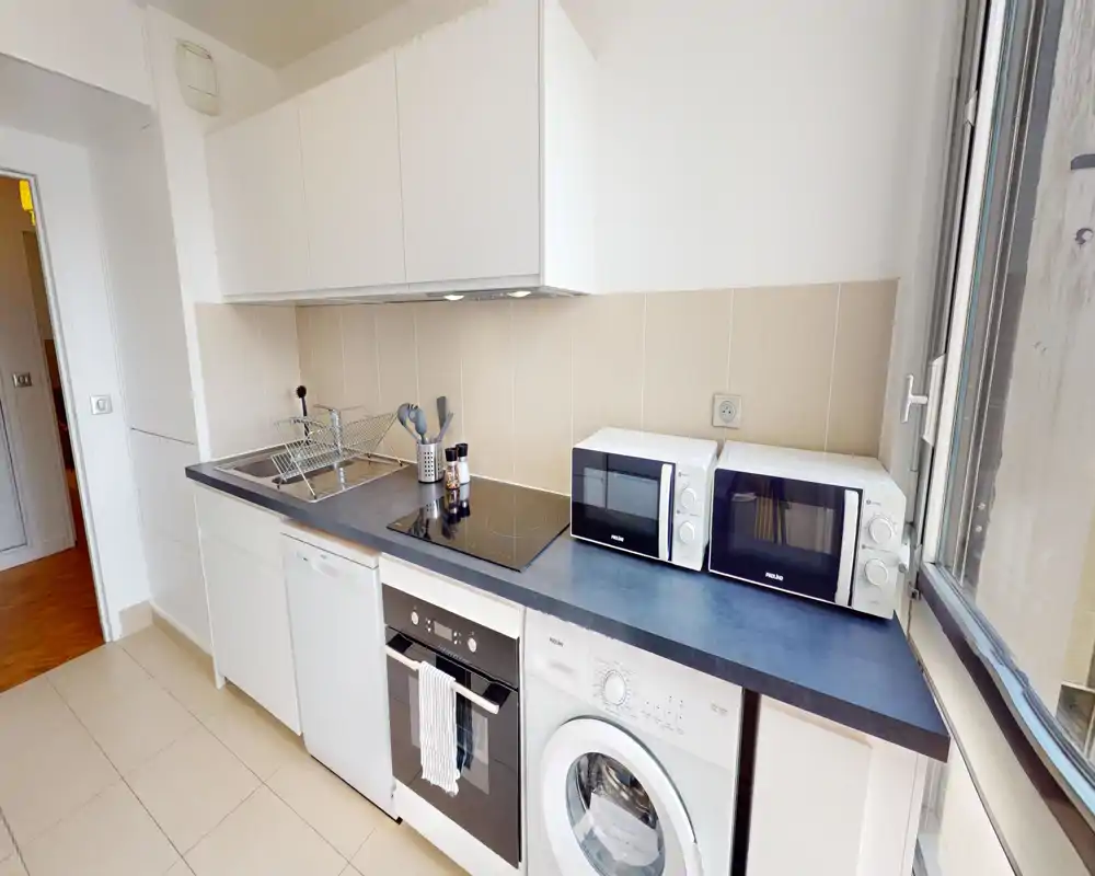 Modern compact kitchen with integrated appliances, ceramic hob, countertop microwave ovens and a washing machine beneath the counter. Large window provides natural light and the layout is tidy and functional — ideal for short-term rentals.