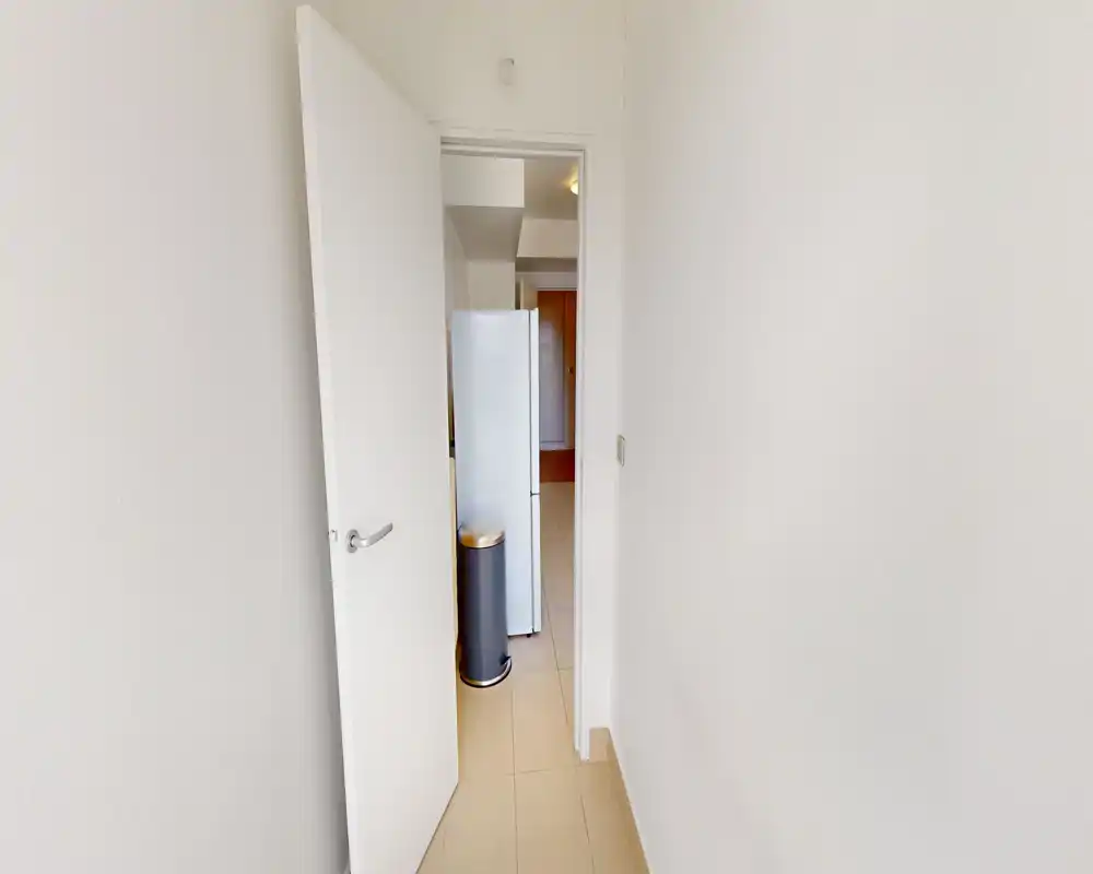 Narrow entry corridor with a partially open white door leading into the apartment; tiled floor and neutral white walls create a clean, minimal entrance area.