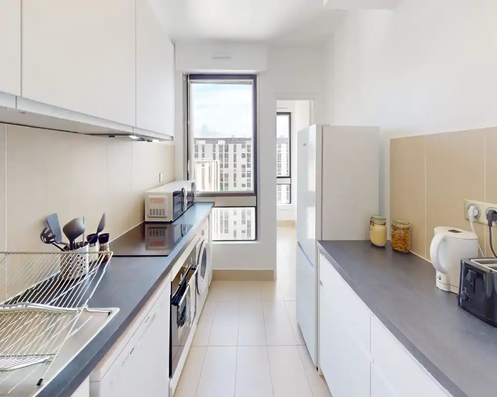 Bright modern galley kitchen with white cabinets, dark countertops, built-in oven, refrigerator and windowed view — clean and functional space ideal for apartment listings.