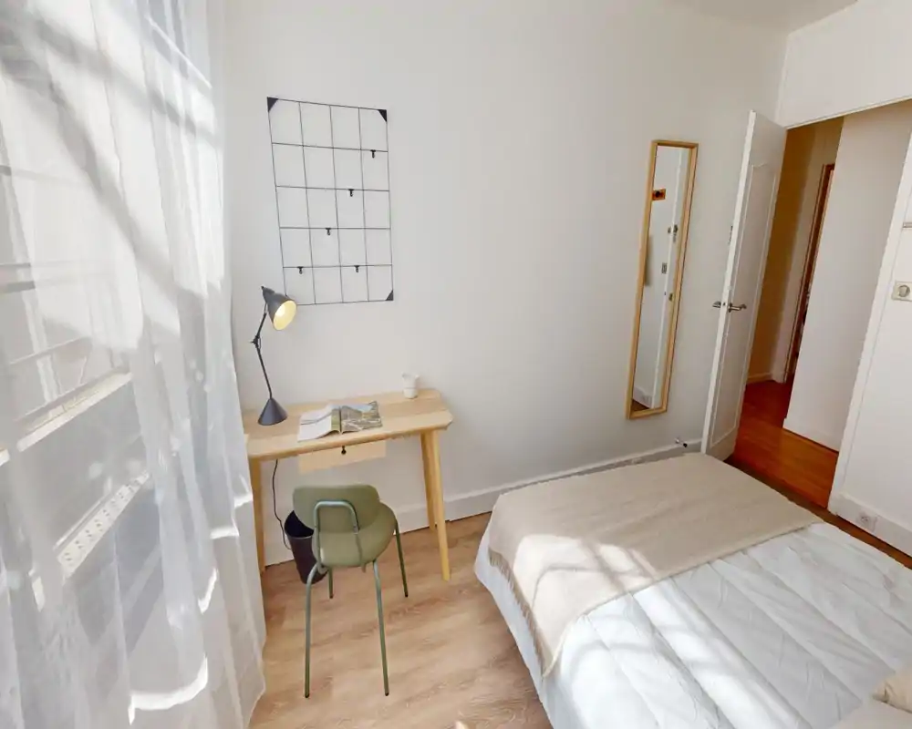 Chambre lumineuse et minimaliste avec un lit confortable, une grande fenêtre laissant entrer la lumière naturelle et un petit bureau en bois—parfait pour travailler ou se détendre.