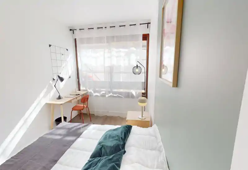 Bright, minimal bedroom with a comfortable double bed, bedside table and a desk by a large window that fills the room with natural light.