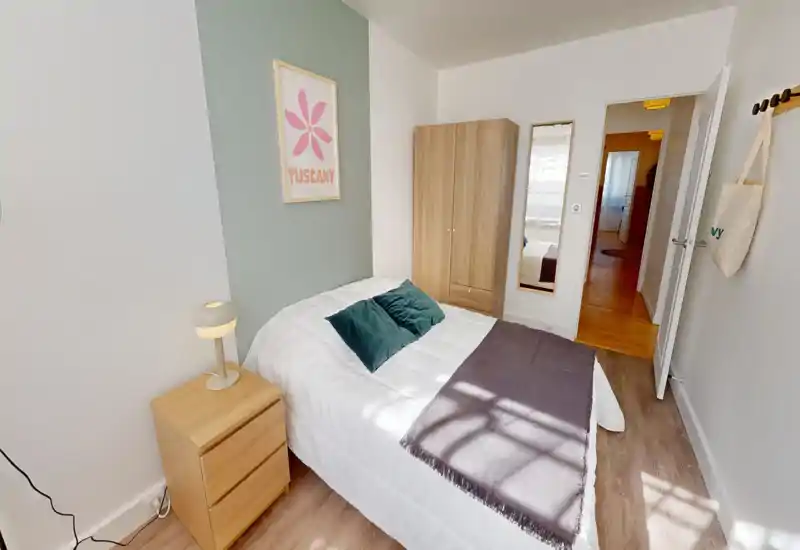 Bright, cozy bedroom with a neatly made double bed, green accent wall and matching wood furniture — bedside table and wardrobe. Natural light and tasteful cushions create a welcoming atmosphere.