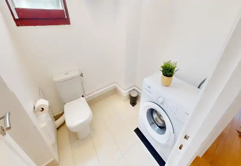 Salle de bain compacte avec toilette et lave-linge à hublot, sol carrelé et une petite fenêtre dépoli apportant de la lumière naturelle.