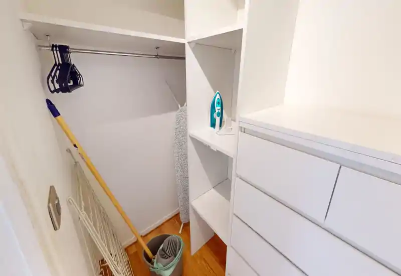 Petit dressing/rangement d'entrée avec étagères blanches intégrées, tiroirs et tringle. On y voit des ustensiles de nettoyage, un fer à repasser et une planche : espace lumineux et fonctionnel pour organiser vêtements et affaires.