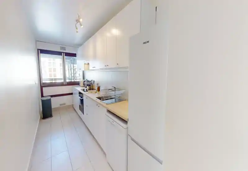 Cuisine en longueur moderne et lumineuse avec placards blancs, plan de travail en bois et électroménagers intégrés. Grande fenêtre et surfaces propres pour un espace fonctionnel.