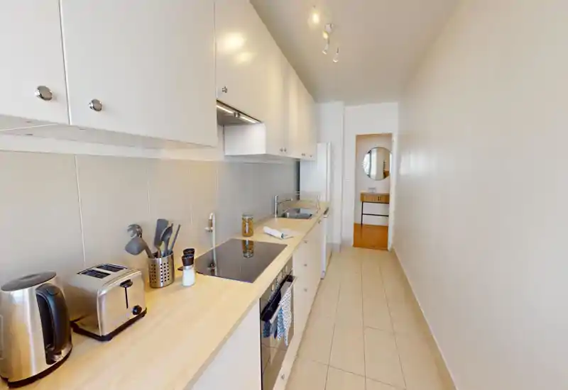 Cuisine moderne et étroite avec plan de travail en bois clair, placards blancs, plaque à induction, grille-pain et bouilloire ; lumineuse et soigneusement mise en scène.
