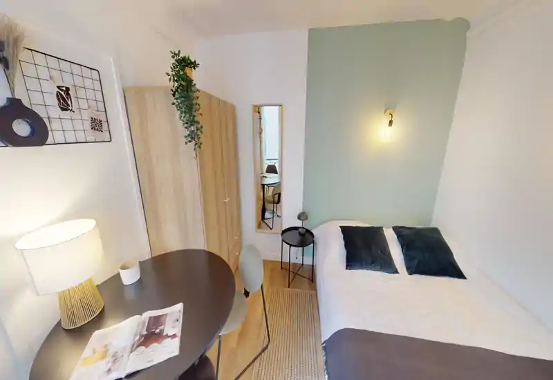 Chambre simple lumineuse et chaleureuse avec lit bien fait, table de chevet et petit coin bureau — idéale pour un étudiant ou voyageur seul.