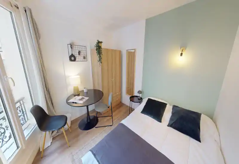 Chambre lumineuse et compacte avec un lit double, textiles doux et une petite table près de la fenêtre ; armoire en bois clair et lampe murale pour une ambiance chaleureuse et moderne.