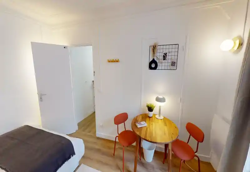 Coin chambre chaleureux et bien éclairé avec lit visible, petite table ronde en bois et deux chaises — parfait pour une location compacte. Murs blancs, lampe et sol en bois apportent une ambiance accueillante.