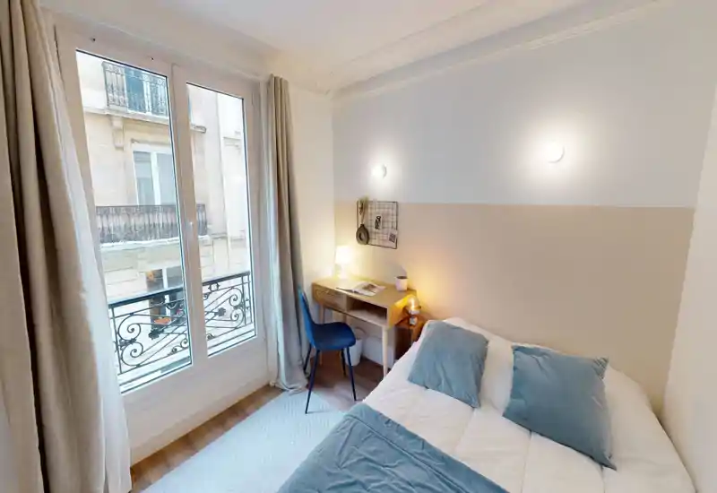 Chambre lumineuse et cosy avec lit double, oreillers moelleux et bureau de chevet sous des appliques chaleureuses ; grande fenêtre avec balcon apporte beaucoup de lumière naturelle.