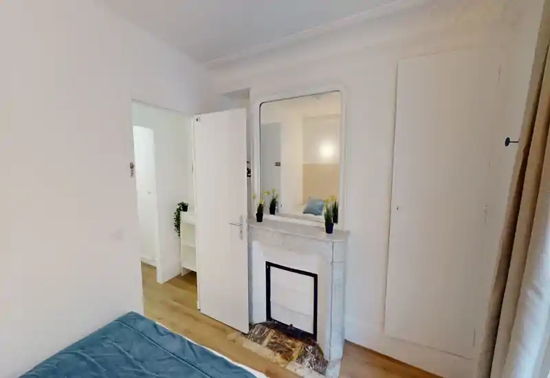 Chambre compacte et lumineuse avec cheminée décorative et grand miroir ; lit soigné avec textiles bleus et décoration sobre pour une ambiance calme.