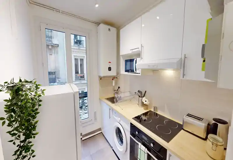 Cuisine moderne et compacte avec placards blancs, plan de travail en bois, plaque à induction, machine à laver et lumière naturelle provenant d'une haute fenêtre — propre et fonctionnelle pour la vie urbaine.