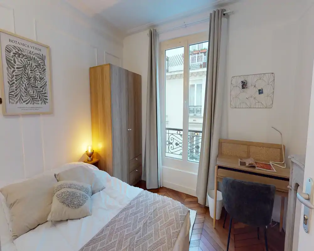 Camera da letto luminosa e accogliente con letto matrimoniale, cuscini decorativi, armadio in legno e una scrivania vicino alla finestra. La luce naturale dalla finestra alta e la lampada sul comodino creano un ambiente caldo e invitante, perfetto per affitti brevi.