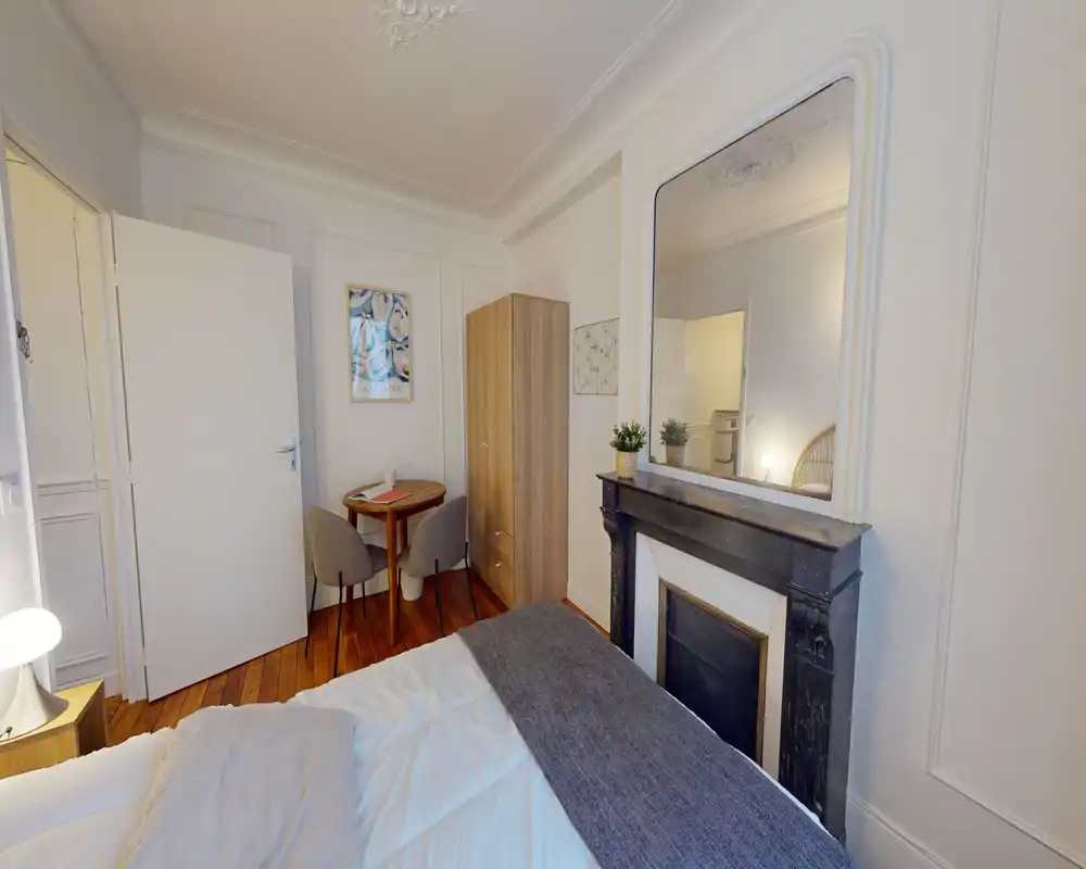 Camera da letto luminosa e accogliente con letto ben rifatto, camino decorativo e grande specchio. Un piccolo tavolo e un armadio sono visibili, ideale per soggiorni brevi.