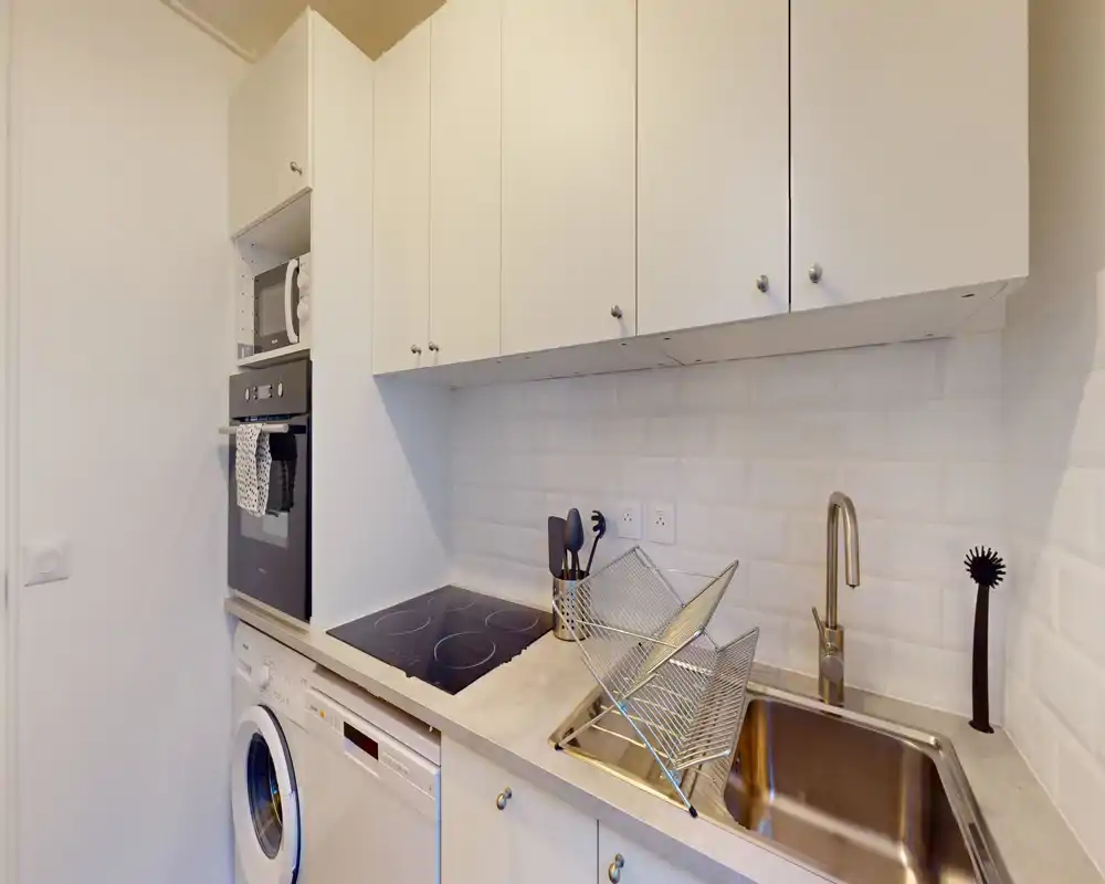 Cocina moderna y compacta con vitrocerámica, horno empotrado, fregadero y lavadora; muebles blancos y salpicadero tipo metro que ofrecen un aspecto limpio y luminoso.