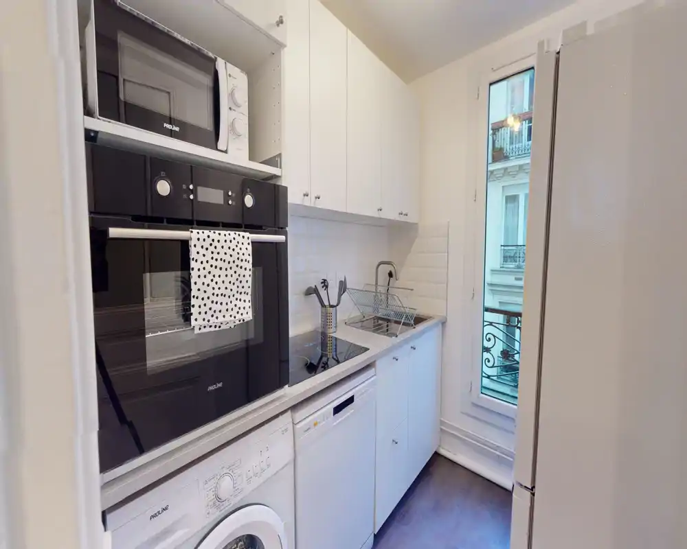 Cocina moderna y compacta con muebles blancos, horno empotrado, microondas y lavadora; la ventana alta aporta luz natural y un diseño limpio y eficiente, ideal para apartamentos en la ciudad.