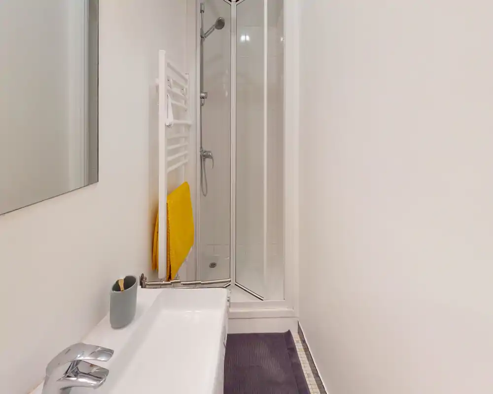 Baño compacto y moderno con plato de ducha en esquina, lavabo blanco y un toallero calefactado con una toalla amarilla.