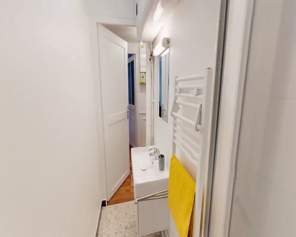 Baño compacto y moderno con pequeño lavabo, espejo, toallero calefactado y una toalla amarilla brillante; paredes blancas y suelo de mosaico para un aspecto limpio y minimalista.