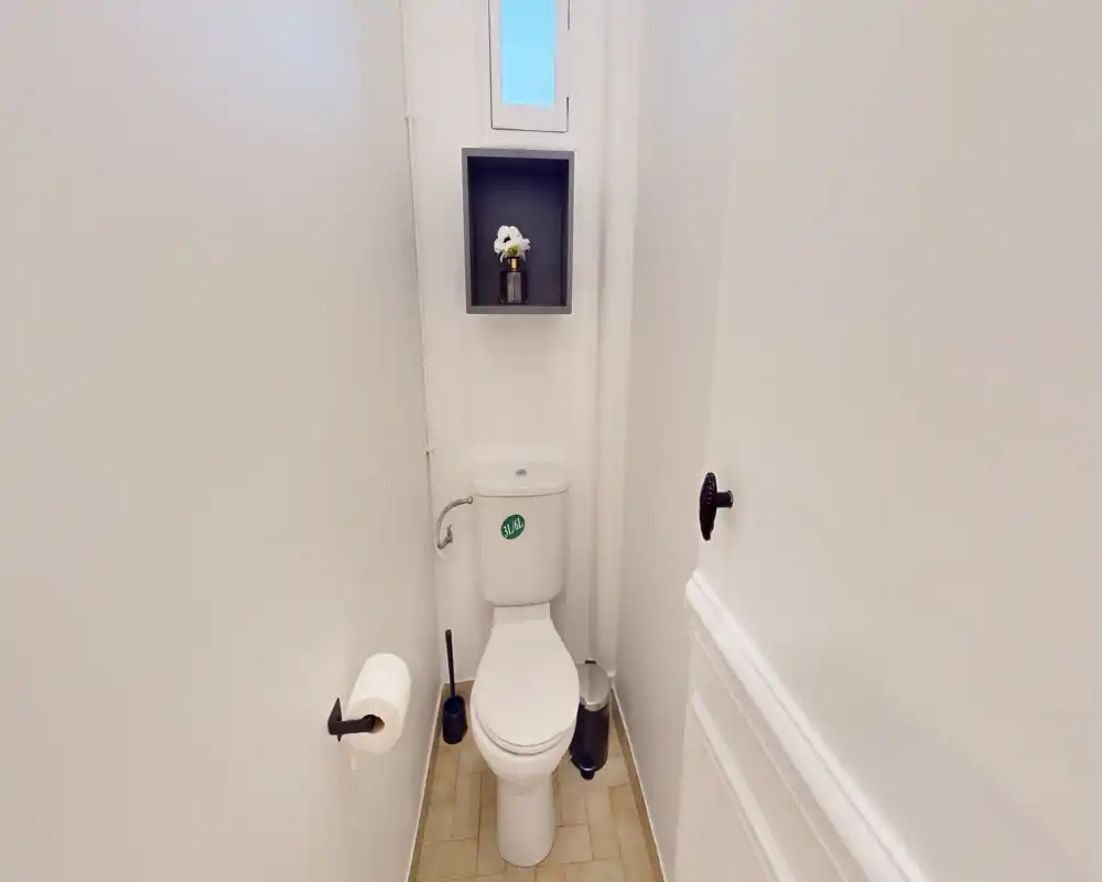 Aseo compacto y luminoso con WC de suelo, escobilla y estante discreto sobre el depósito. Paredes blancas y suelo de baldosa para un aseo de invitados minimalista y bien mantenido.