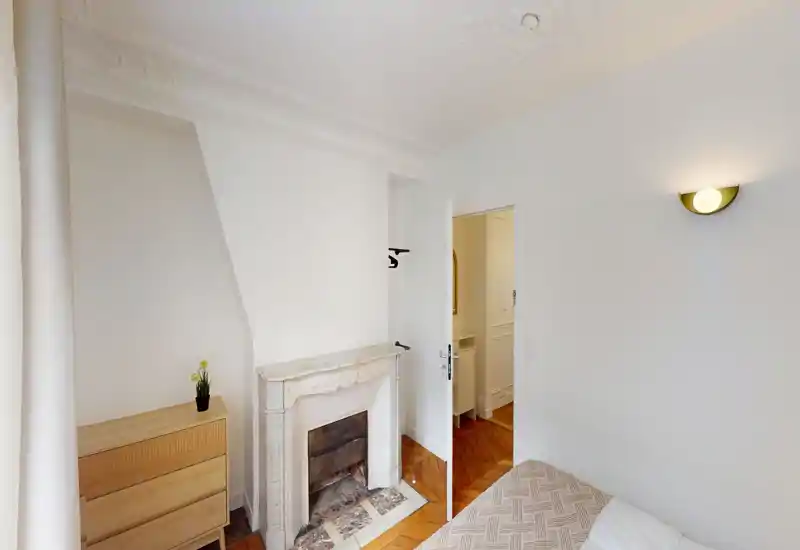 Coin de chambre lumineux avec une cheminée en marbre décorative, parquet et lit partiellement visible. Murs blancs et lumière naturelle créent une ambiance aérée et minimaliste.