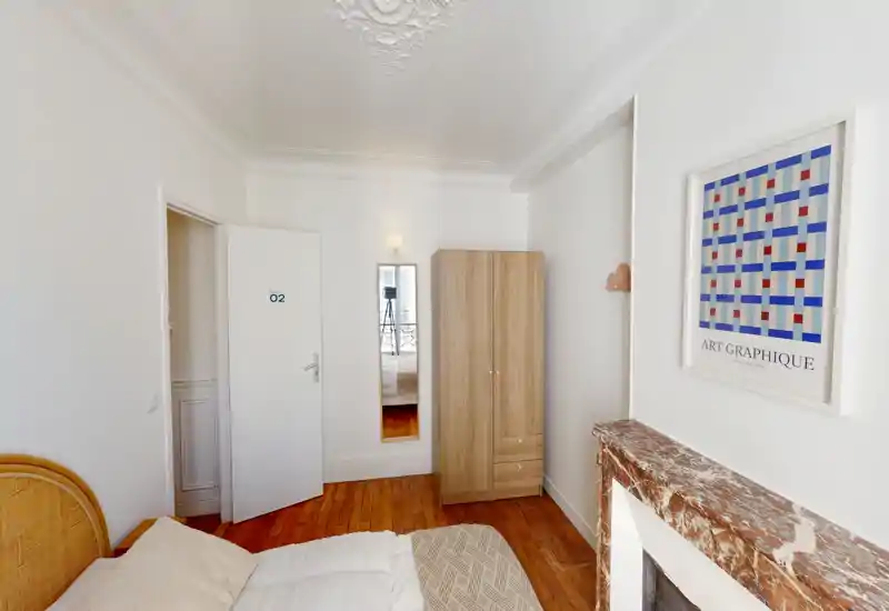 Chambre lumineuse et simple avec vue partielle du lit, parquet, armoire et cheminée décorative ; murs blancs et cadre apportent une atmosphère aérée.