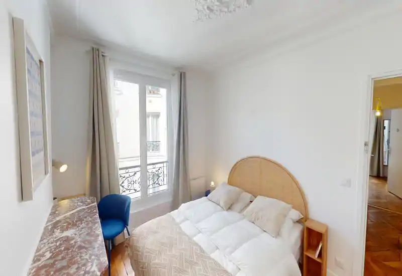 Chambre lumineuse et soignée avec tête de lit en rotin, literie impeccable et fenêtre apportant une belle lumière naturelle ; petit bureau en marbre et chaise bleue.
