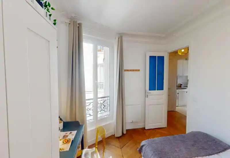 Chambre lumineuse et chaleureuse avec parquet, grande fenêtre et moulures élégantes — coin bureau pratique et lit partiellement visible avec textiles confortables.